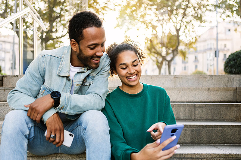 Two young latin american people using smart phone together sitting outdoors Social + Digital Media<br />